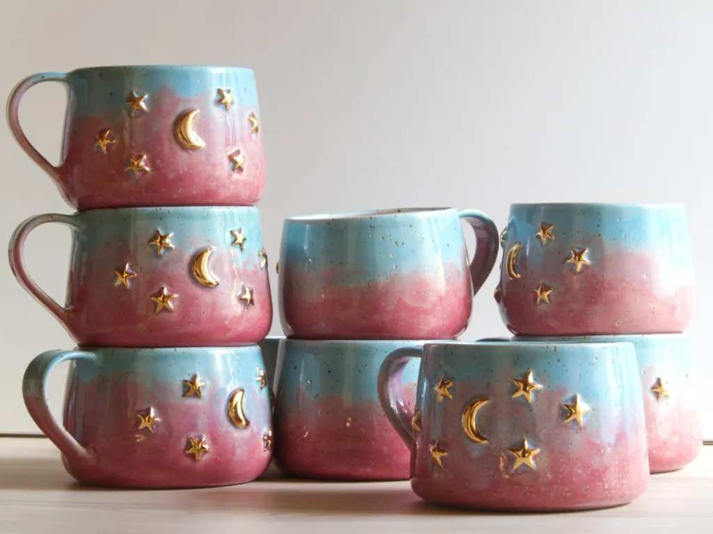 A collection of handmade stoneware mugs with a blue and pink gradient glaze, decorated with hand-applied genuine gold stars and crescent moons.