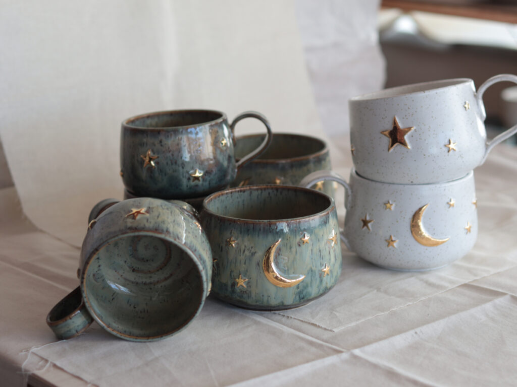 Handmade Celestial Mugs in green and white glazes by artisan Seyma Ates. These unique stoneware mugs feature intricate real gold details of crescent moons and stars, presented on a rustic linen background