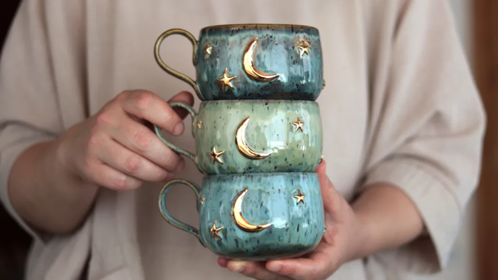 A stack of three handmade stoneware mugs from the Celestial series featuring teal and turquoise glazes with genuine gold luster moon and star details, held by the artist